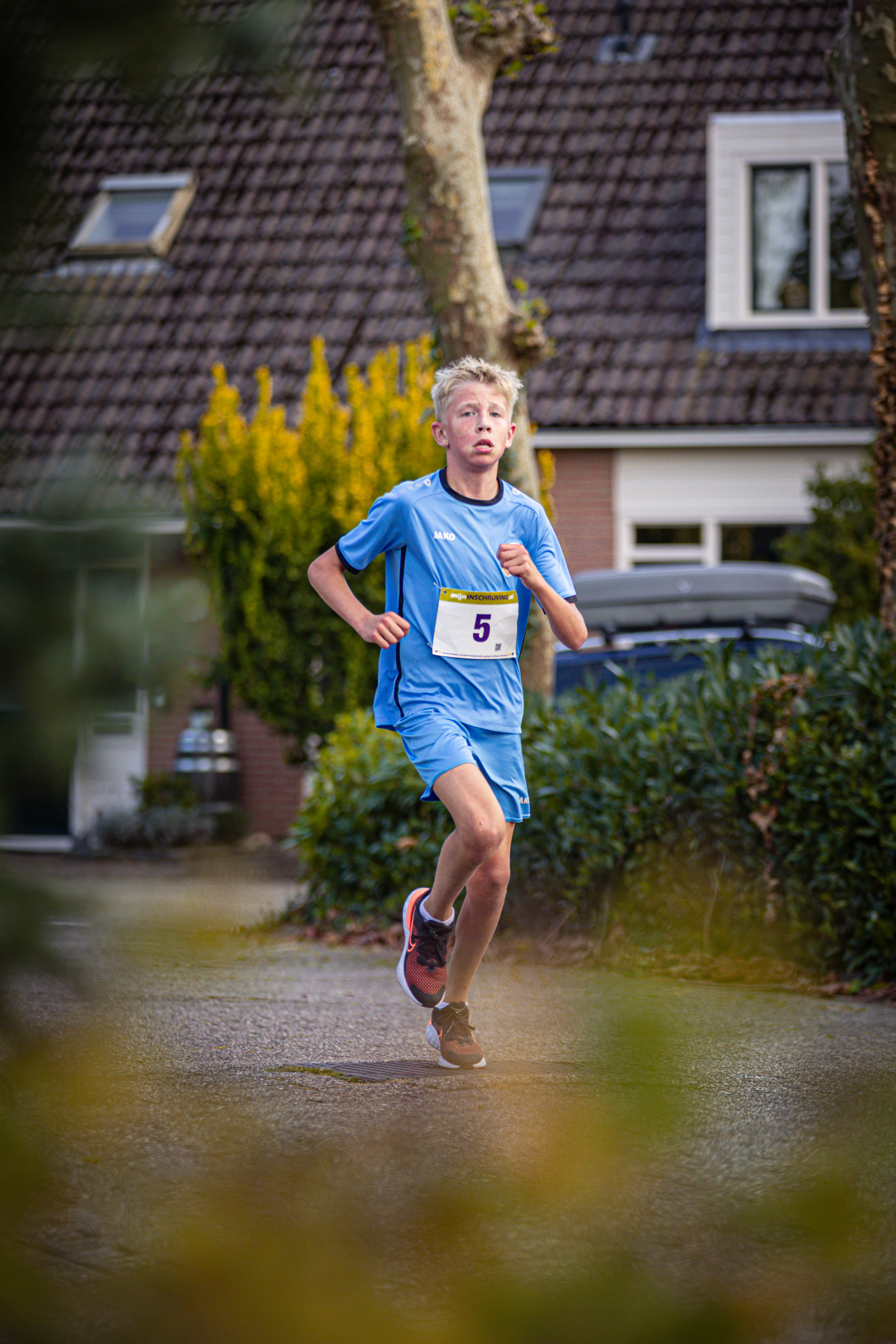 A boy in a blue running outfit with the number 5 on it is running on a wet street.