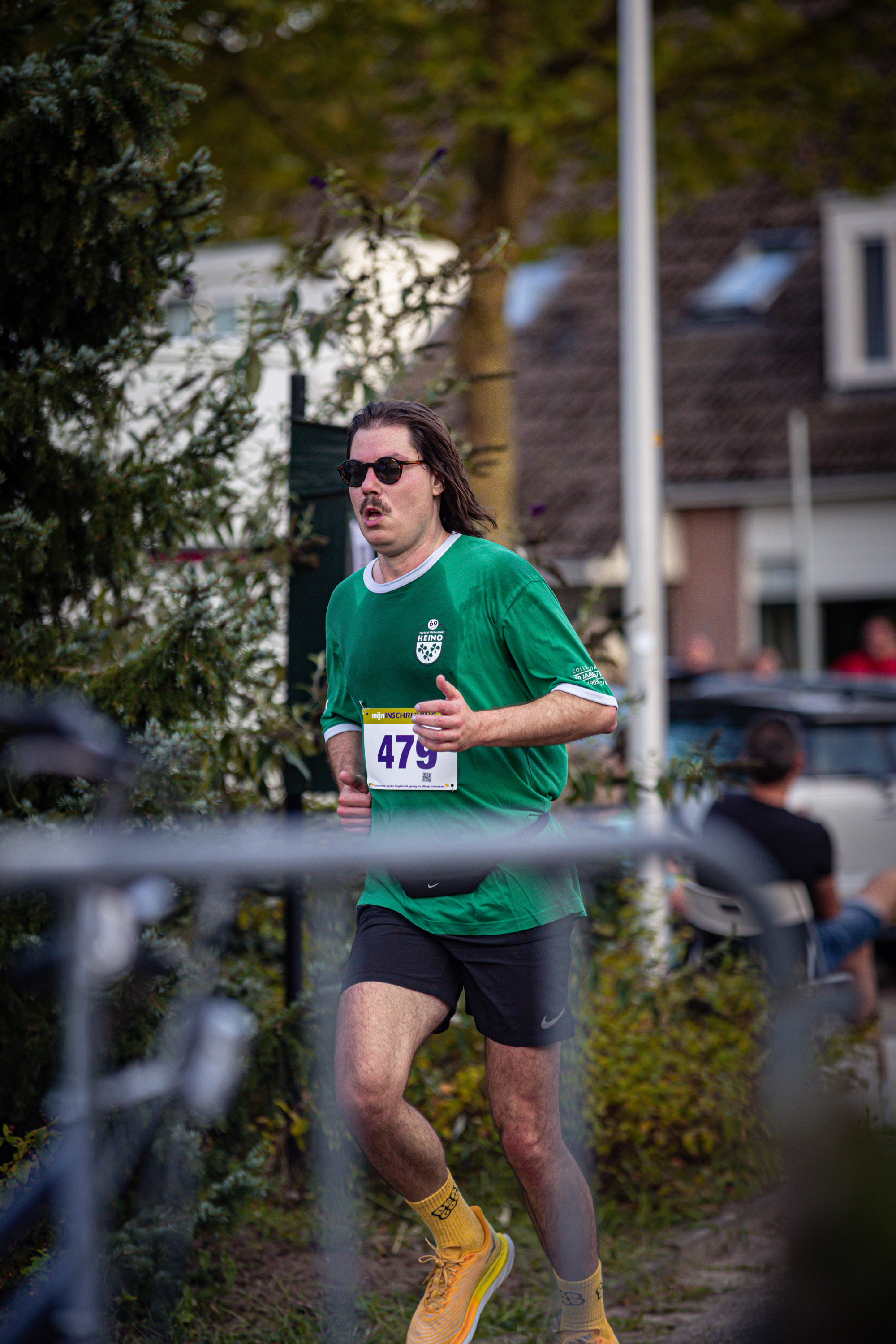 A man running with a green shirt that says Pomp on it. He is wearing black shorts and yellow shoes. His bib number is 475.