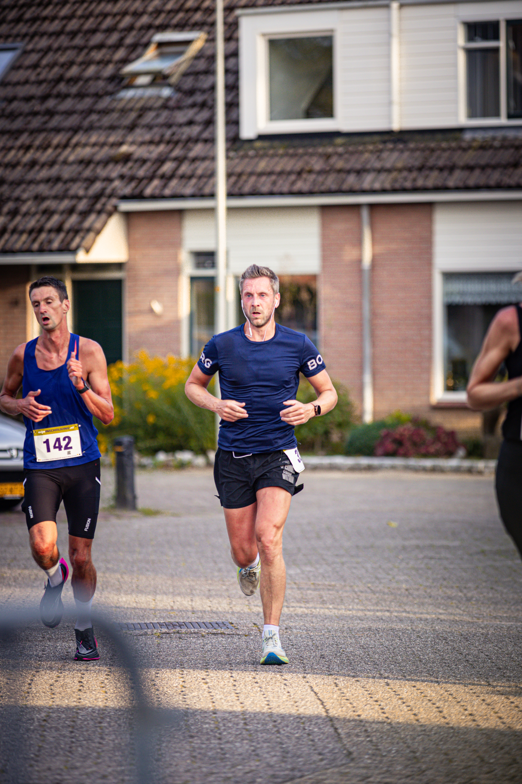 Two runners, one in a blue shirt with the number 145 and the other wearing black, are running past a building.