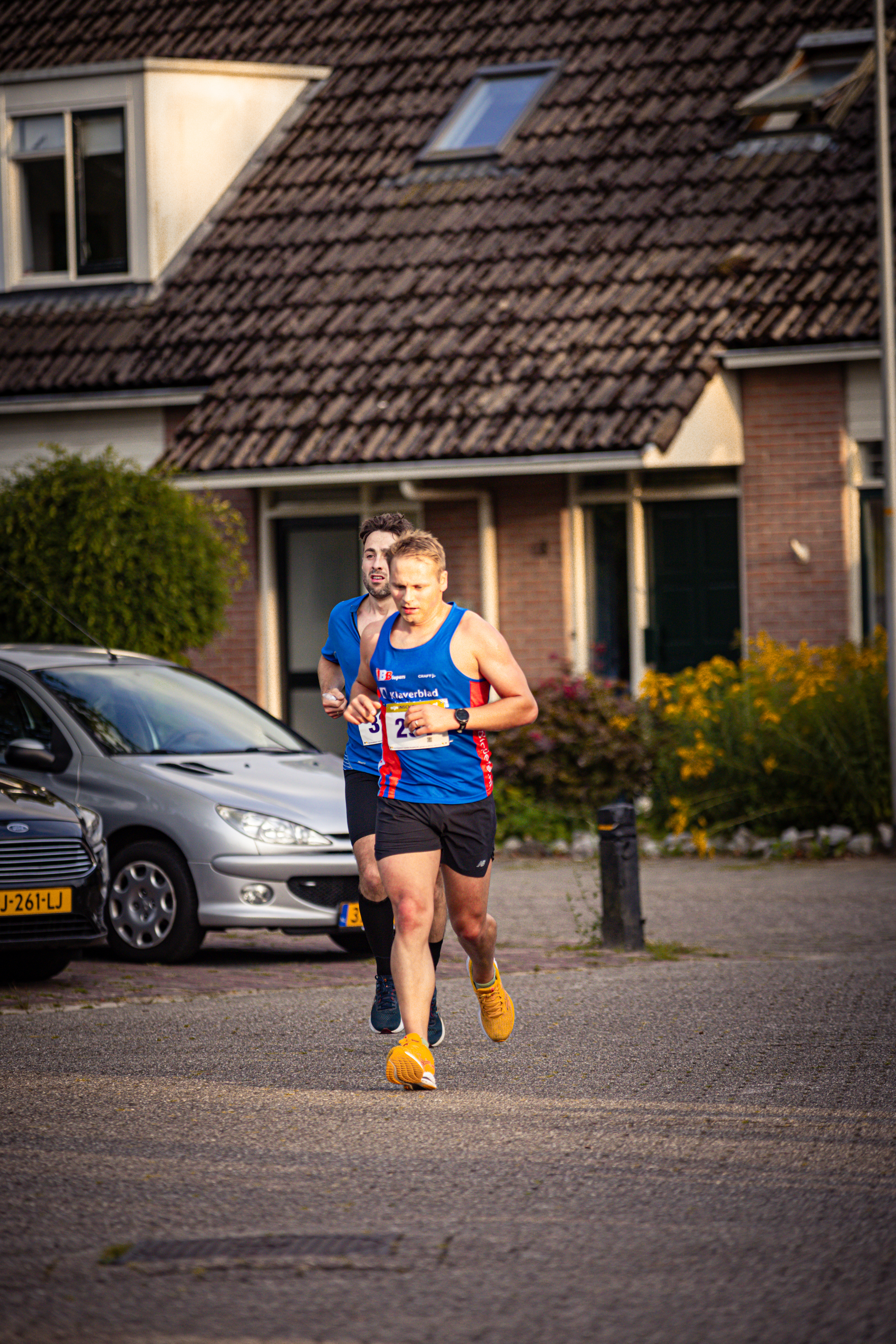 A man and a woman jogging in front of a house with a red car parked nearby.