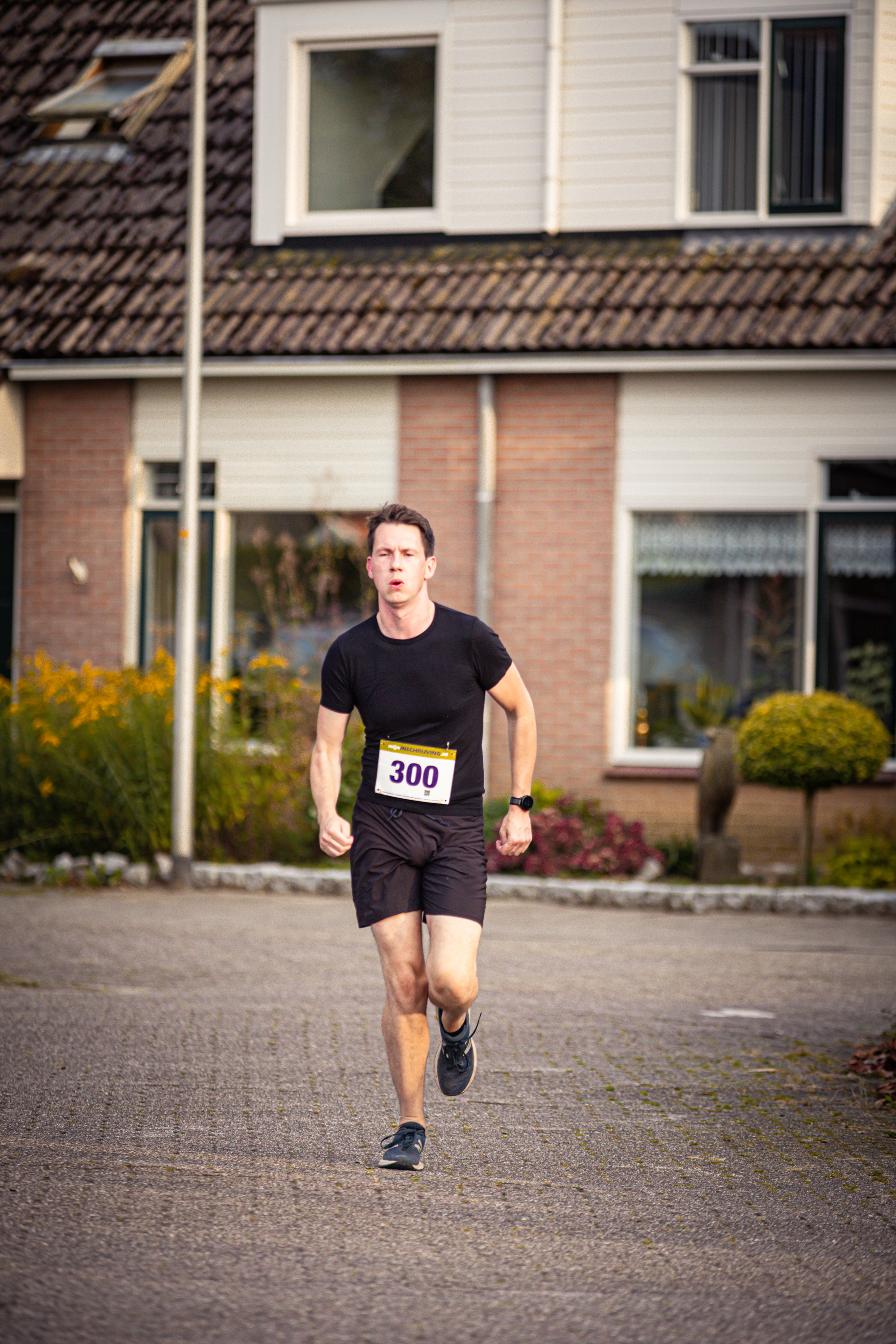 A man wearing a number 300 running jersey runs on the street in front of a white building.