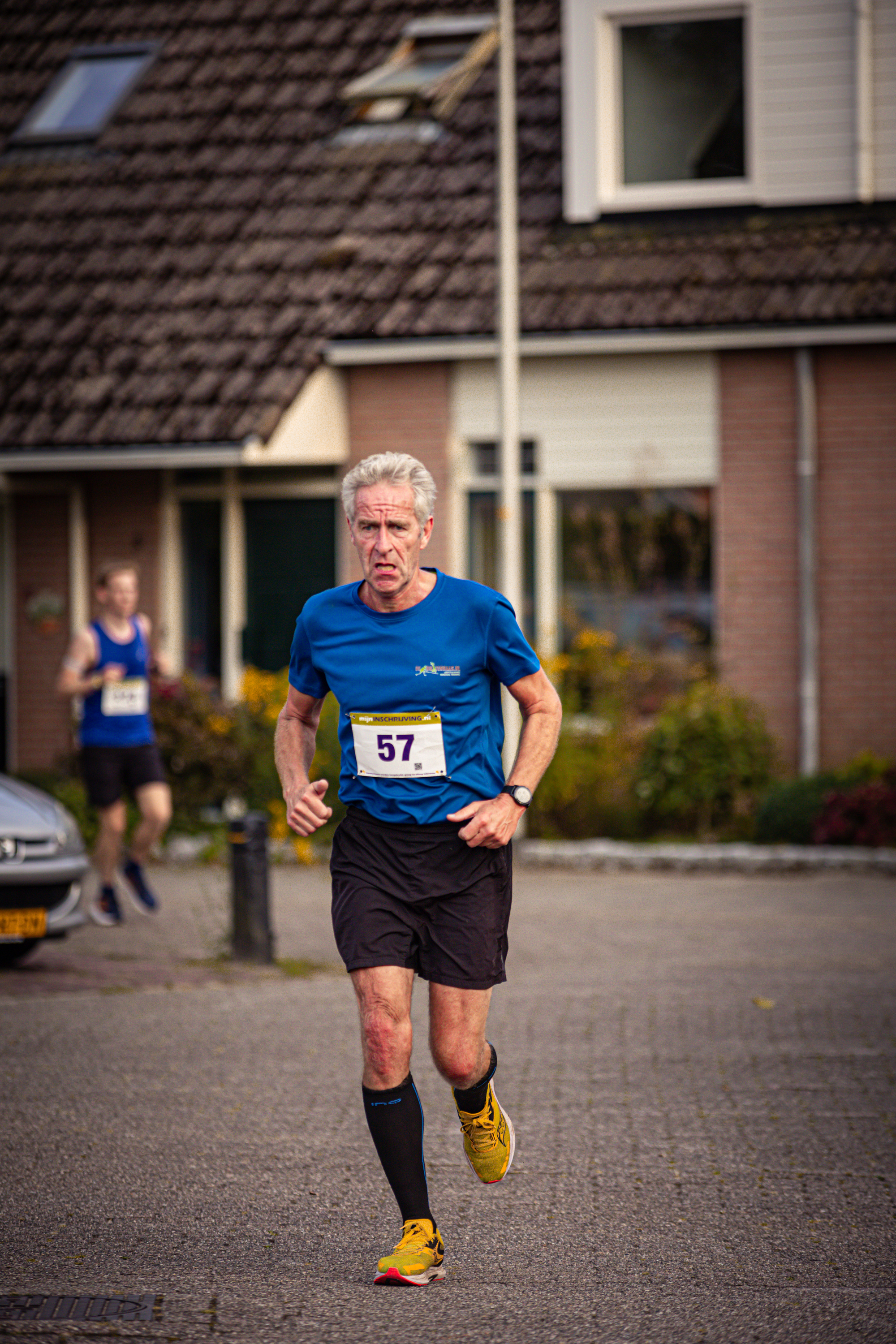 An older man is running in a blue shirt and black shorts. He has the number 57 on his shirt and yellow shoes on his feet.