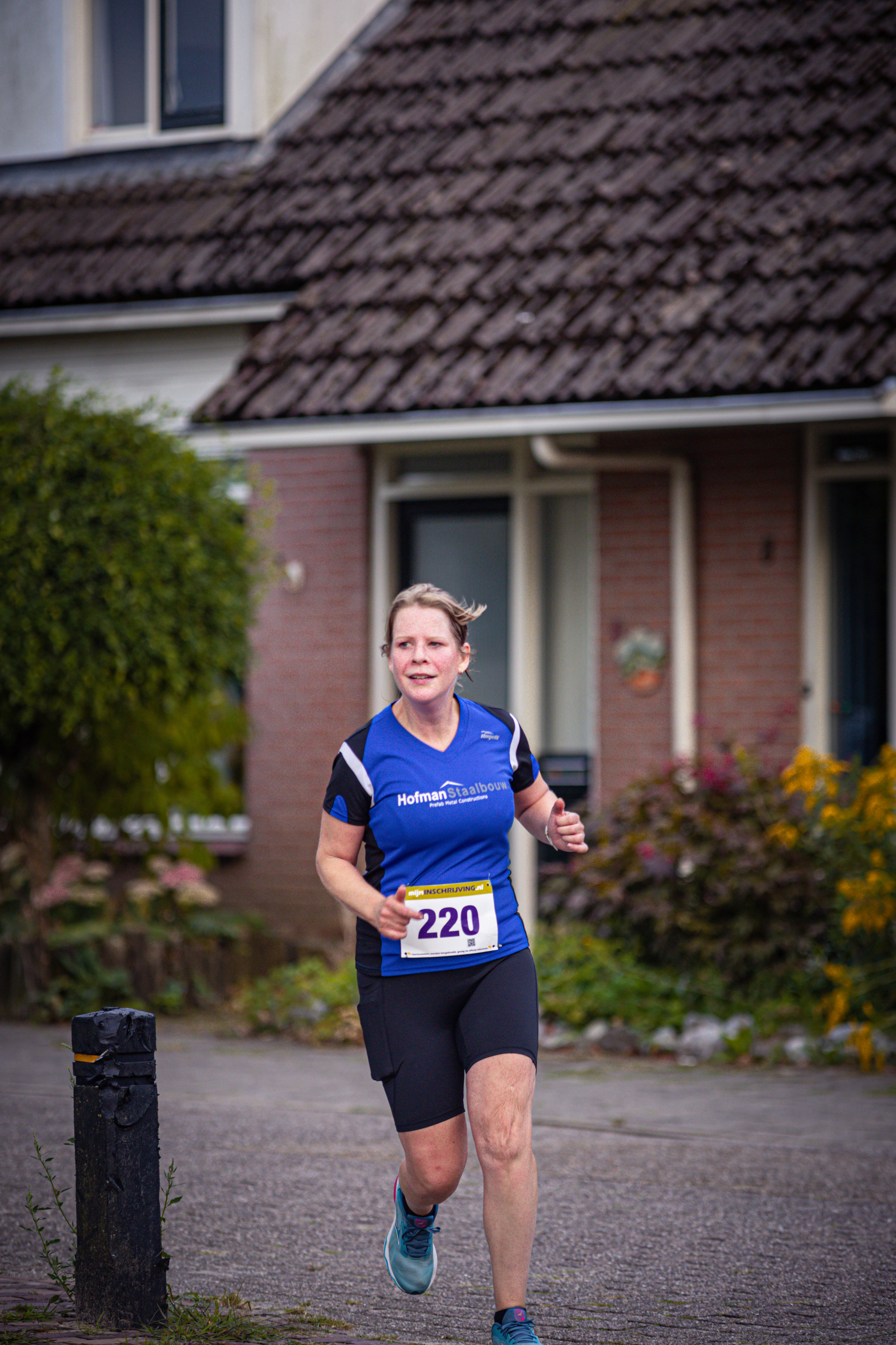 The runner is number 210, running on a sidewalk. She is wearing a blue shirt with the word "Pomp" written on it.