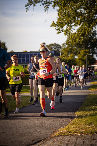 A woman is running on a road with other runners. She has an orange bib and is wearing sunglasses.