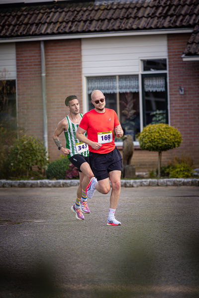 Two men are running on a street. The man in the red shirt is ahead of the other man.