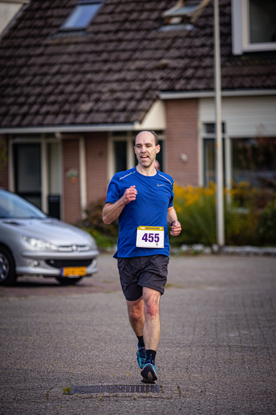 The runner is on the street with a number 459 in his hand. He wears a blue shirt and black shorts and has long hair.