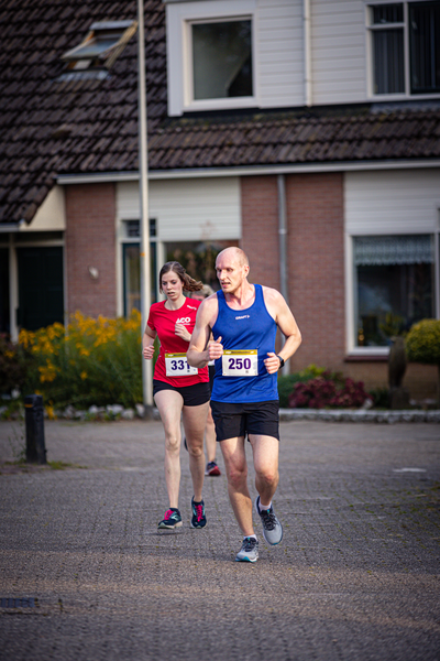 A man and a woman running on a street in front of houses. The man is wearing a shirt that says "Wimblington".