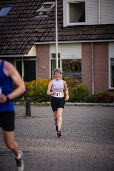 A woman in a purple tank top and black shorts running while wearing a yellow tag that says 21.