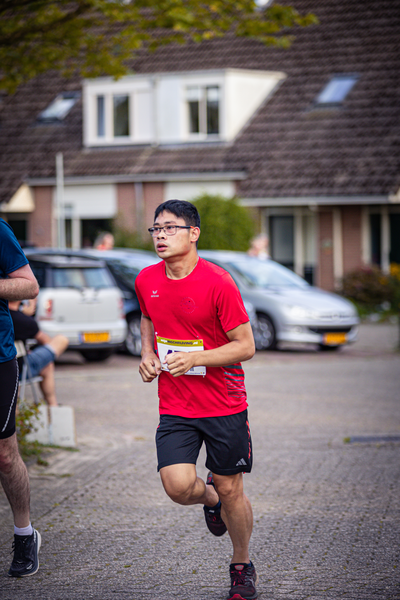 A man wearing a red shirt is running in front of a white car.