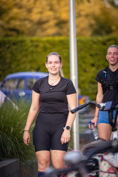 Two women standing in front of a blue car, one is wearing shorts and a black shirt while the other has a black backpack.