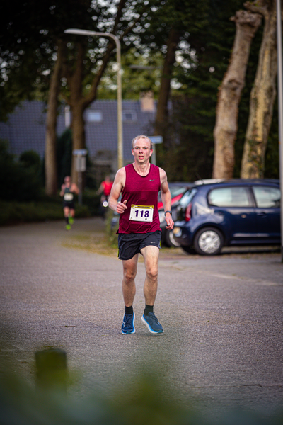 A man running in a red shirt with the number 13 on it. He has blue shoes and is headed toward a car and some trees.