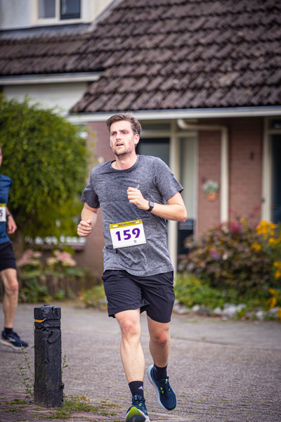 A man in a grey shirt and black shorts runs on the street with an orange number 159 in the middle of his chest.