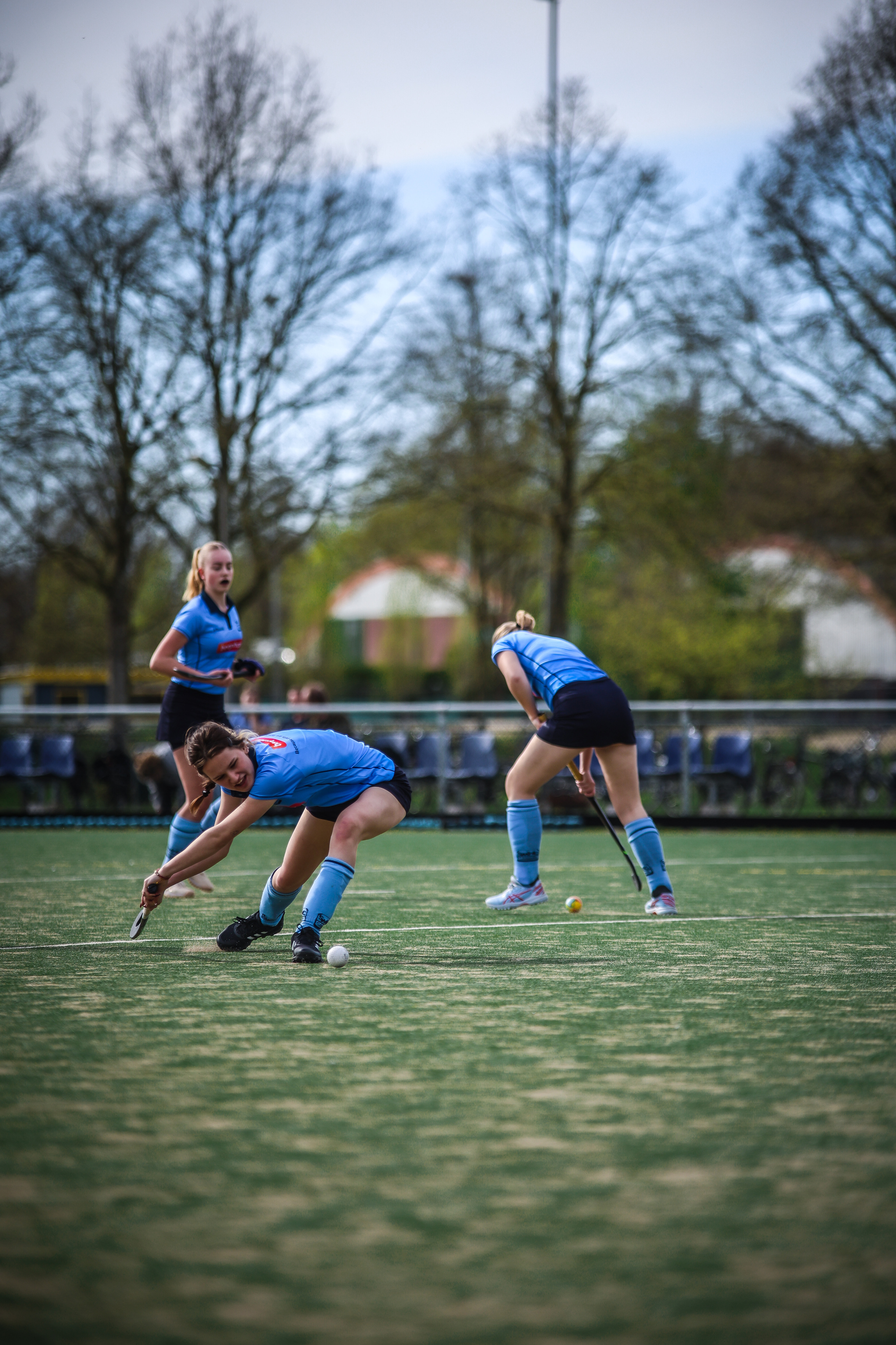 A group of women in blue playing field hockey, one of them is bending over to pick up a ball.