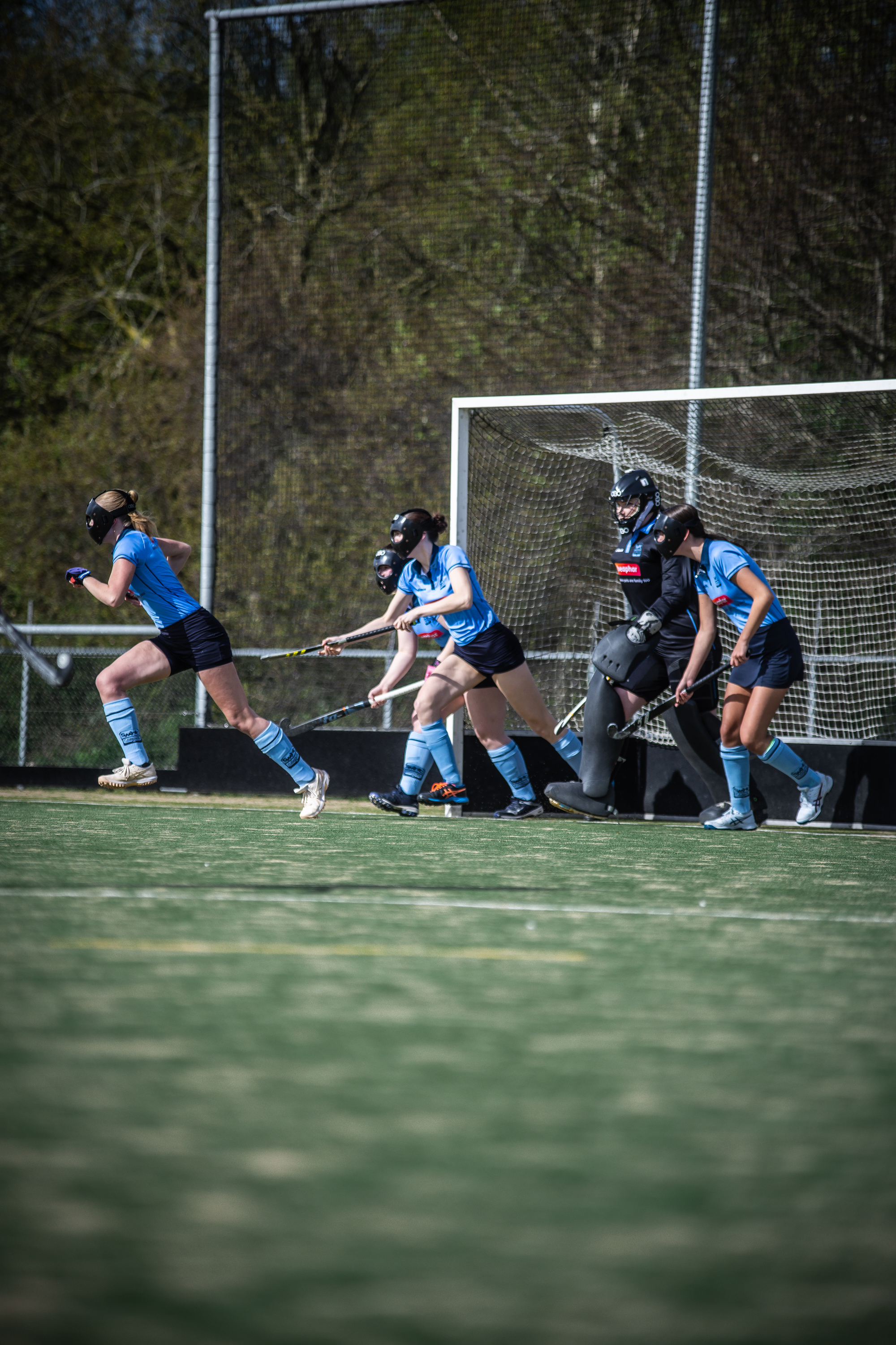 Seven female hockey players in blue uniforms are getting ready to play on a field with a goalie guarding the goal.