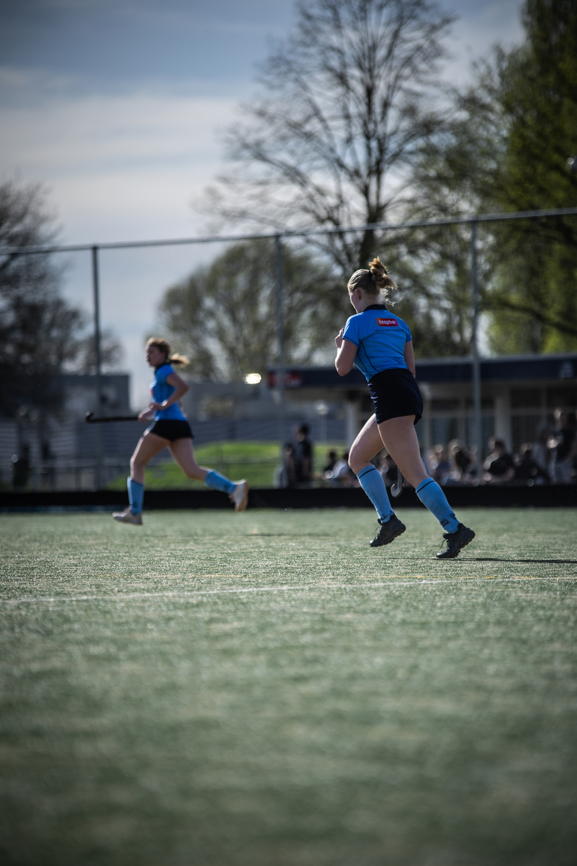 Two girls play hockey on a field with a blue sky and trees in the background.