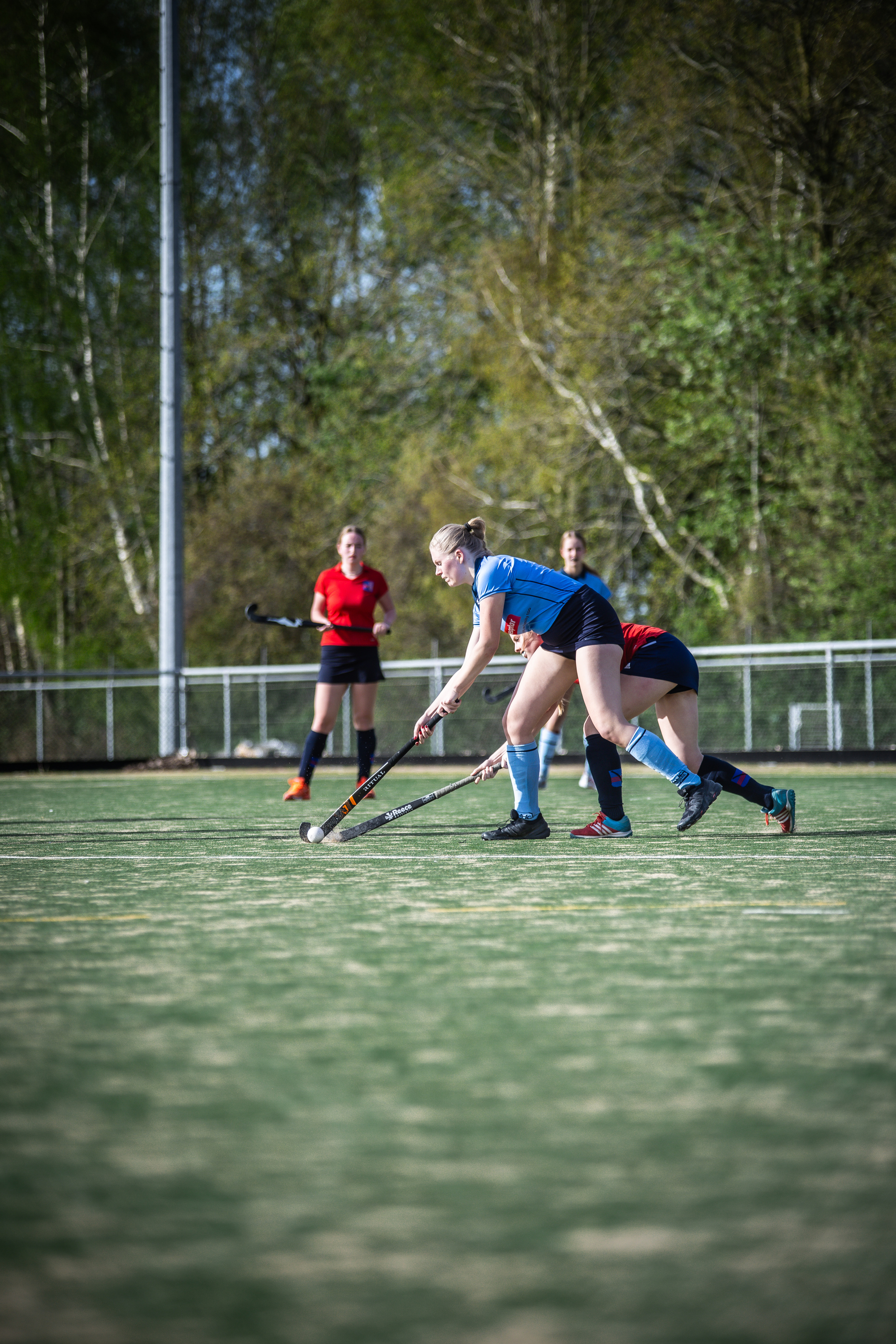 Women's hockey game on a field with green turf and trees in the background.