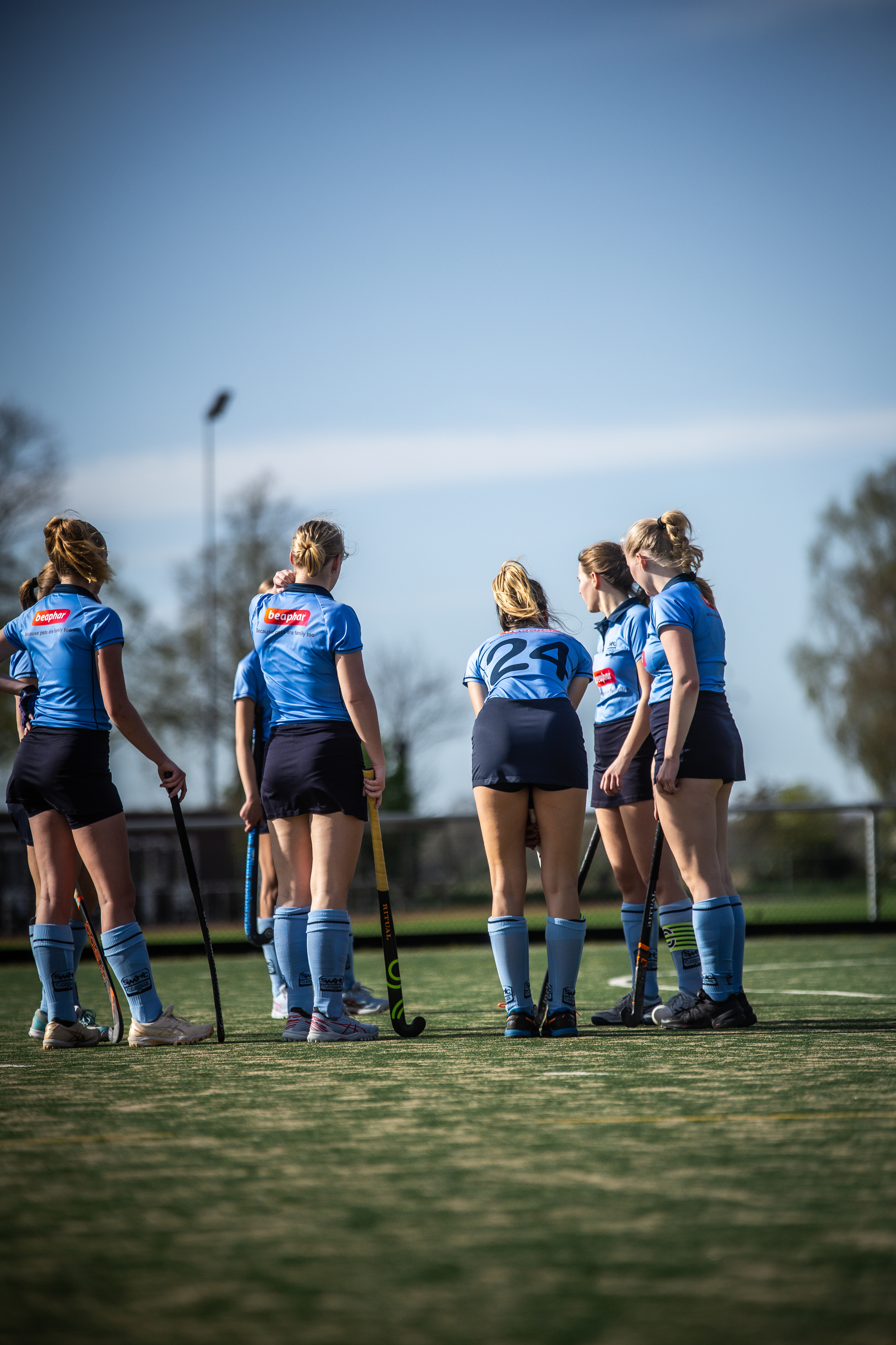 A group of female hockey players stand on a field in blue and white uniforms. They are wearing numbers 2, 3, 4, 6, 7, and 12.