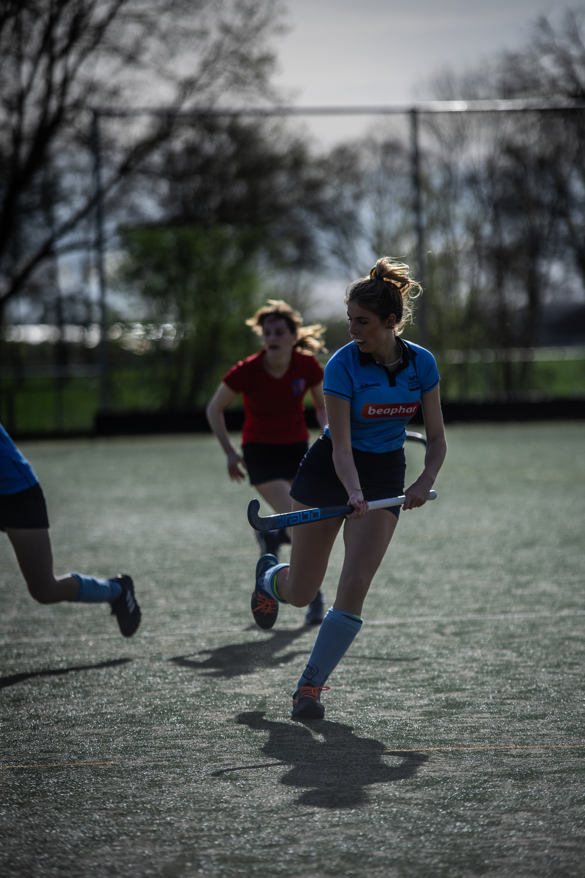 A girl in a blue hockey jersey rushes across the field with her stick in hand.