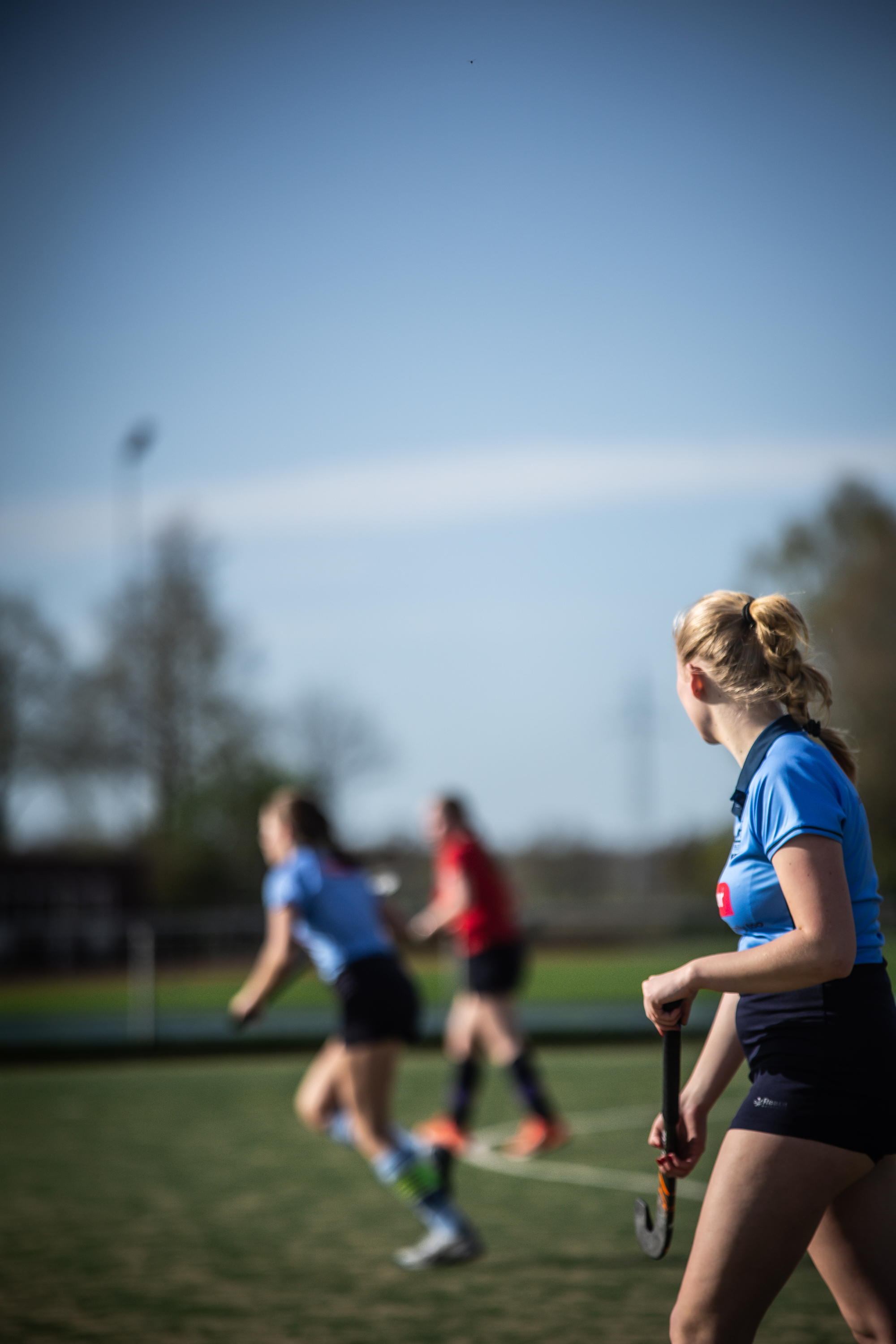 Three girls are playing a game of hockey on a field under a clear blue sky.