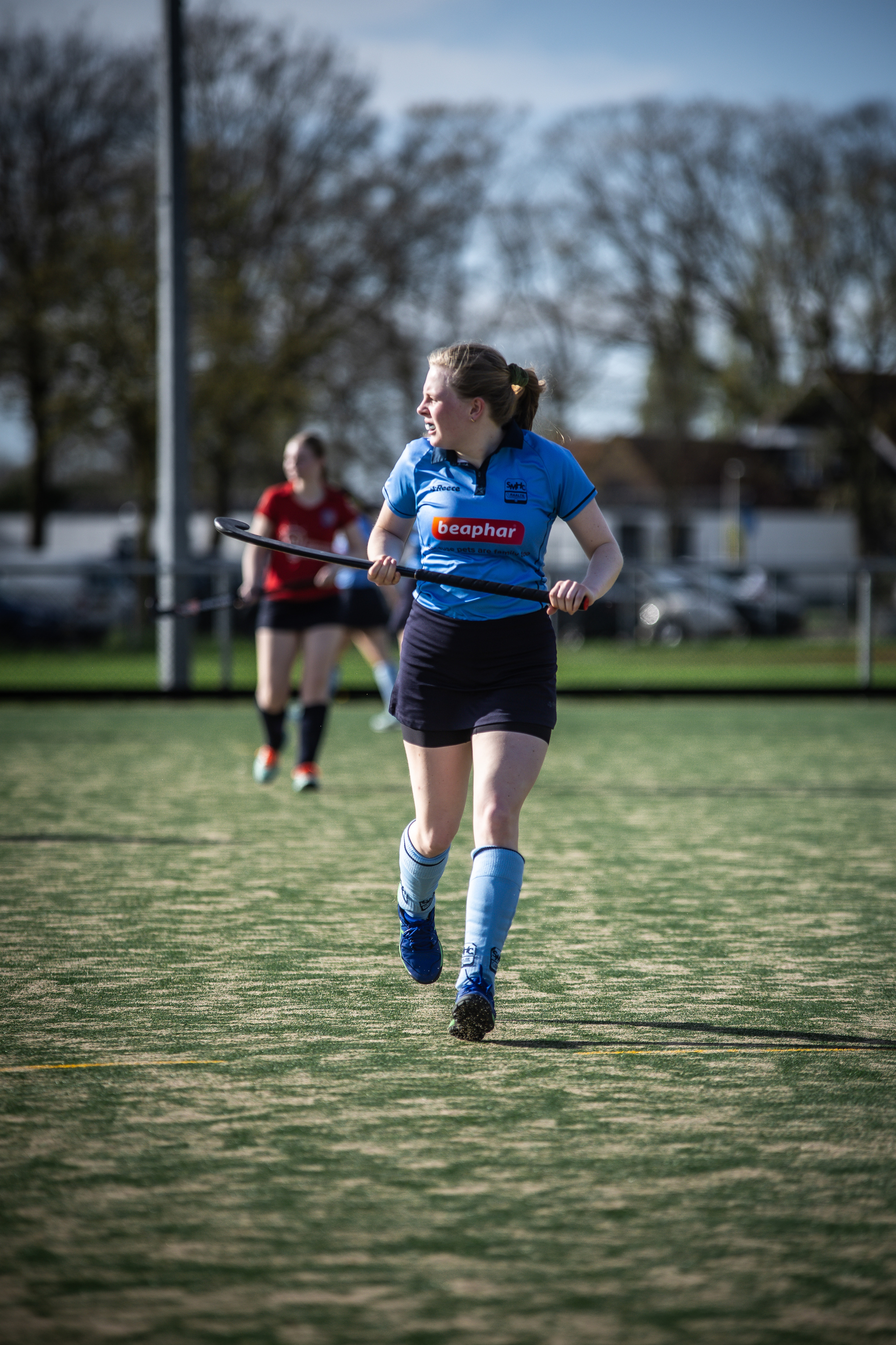 A girl in a blue shirt is playing hockey on an outdoor field. She has her arms outstretched as she runs with the ball.