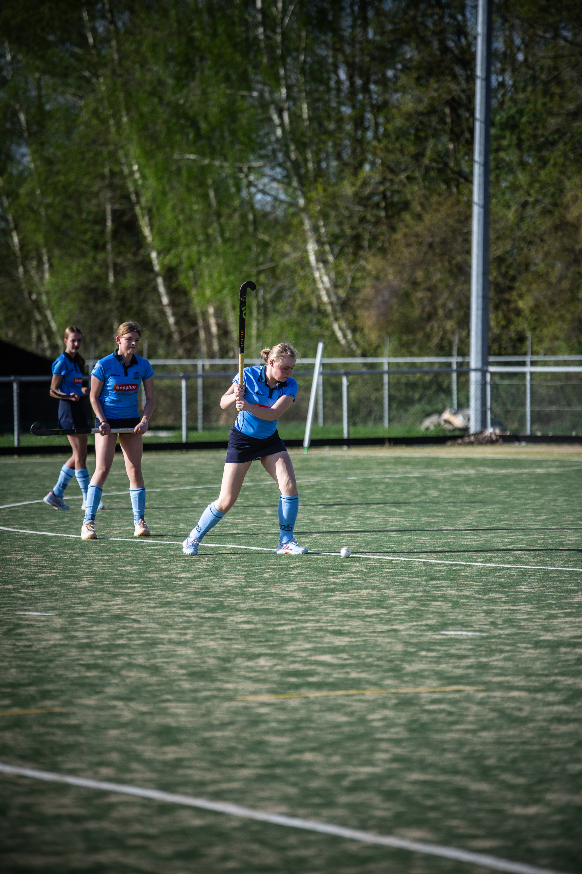 Three women in blue jerseys play hockey on a field.