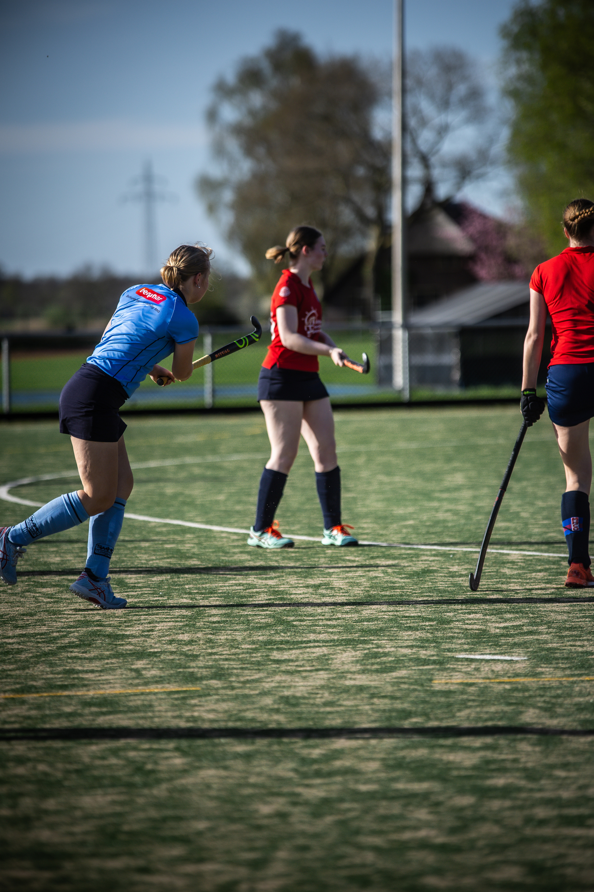 Three women in blue and red uniforms are playing hockey on a field.