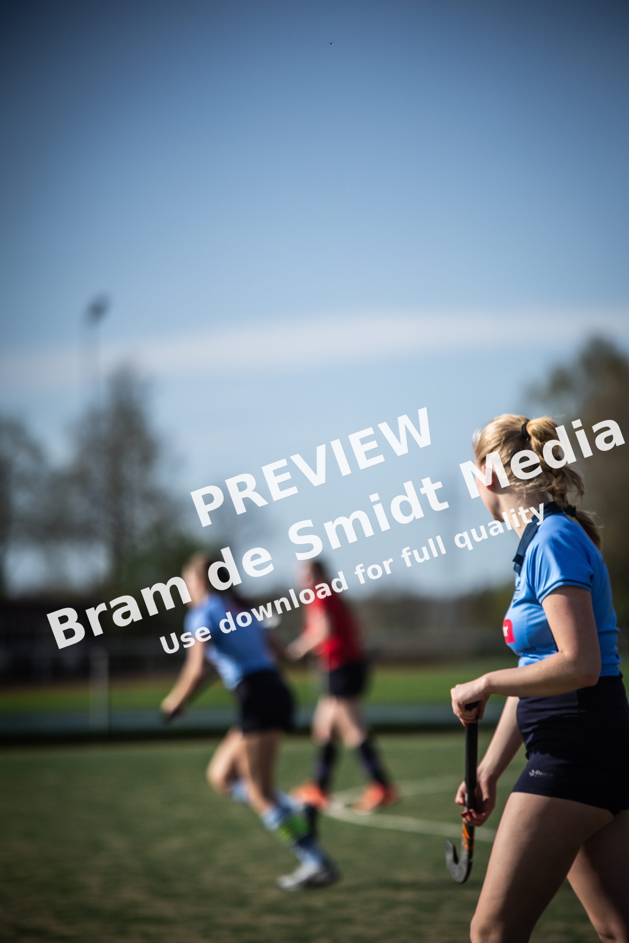 Three girls are playing a game of hockey on a field under a clear blue sky.