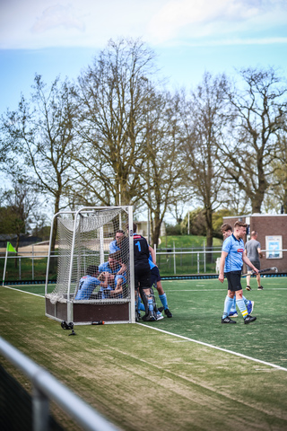 A hockey game in progress, with a goalie guarding the goalpost and players watching the play unfold on a sunny day.