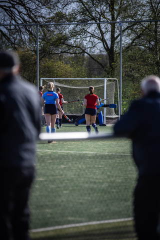 Two girls are playing hockey on a field with a man watching. The girl is wearing a red jersey and the other one is in blue.