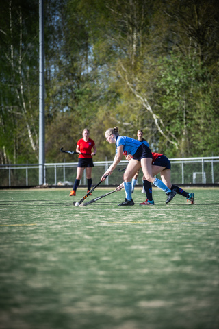 Women's hockey game on a field with green turf and trees in the background.