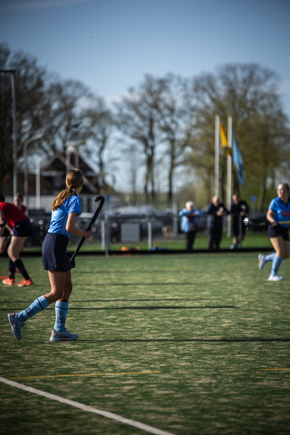 Women play an outdoor game of hockey, the field is marked with lines and there are a few flags in the background.