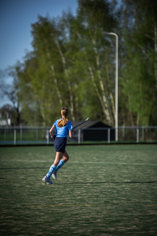 A girl is on a field wearing a blue jersey and shorts, running while holding a hockey stick.