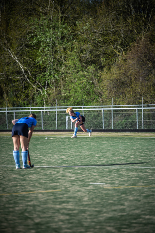 A woman wearing a blue uniform stands on the field with her stick in front of her, ready to hit the ball.