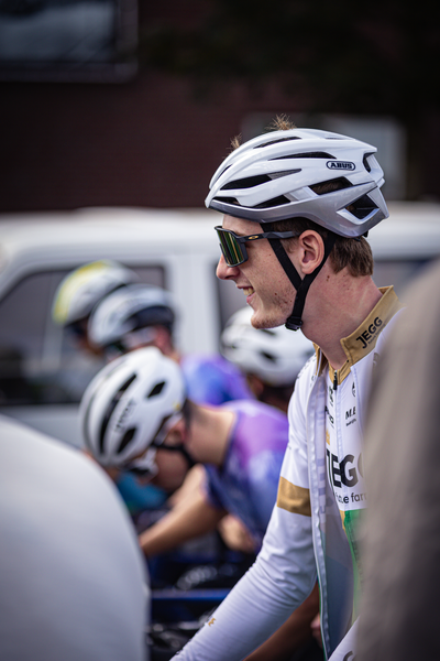 A white man wearing a white bike helmet with the name Giga and blue glasses is riding a bicycle with other cyclists.