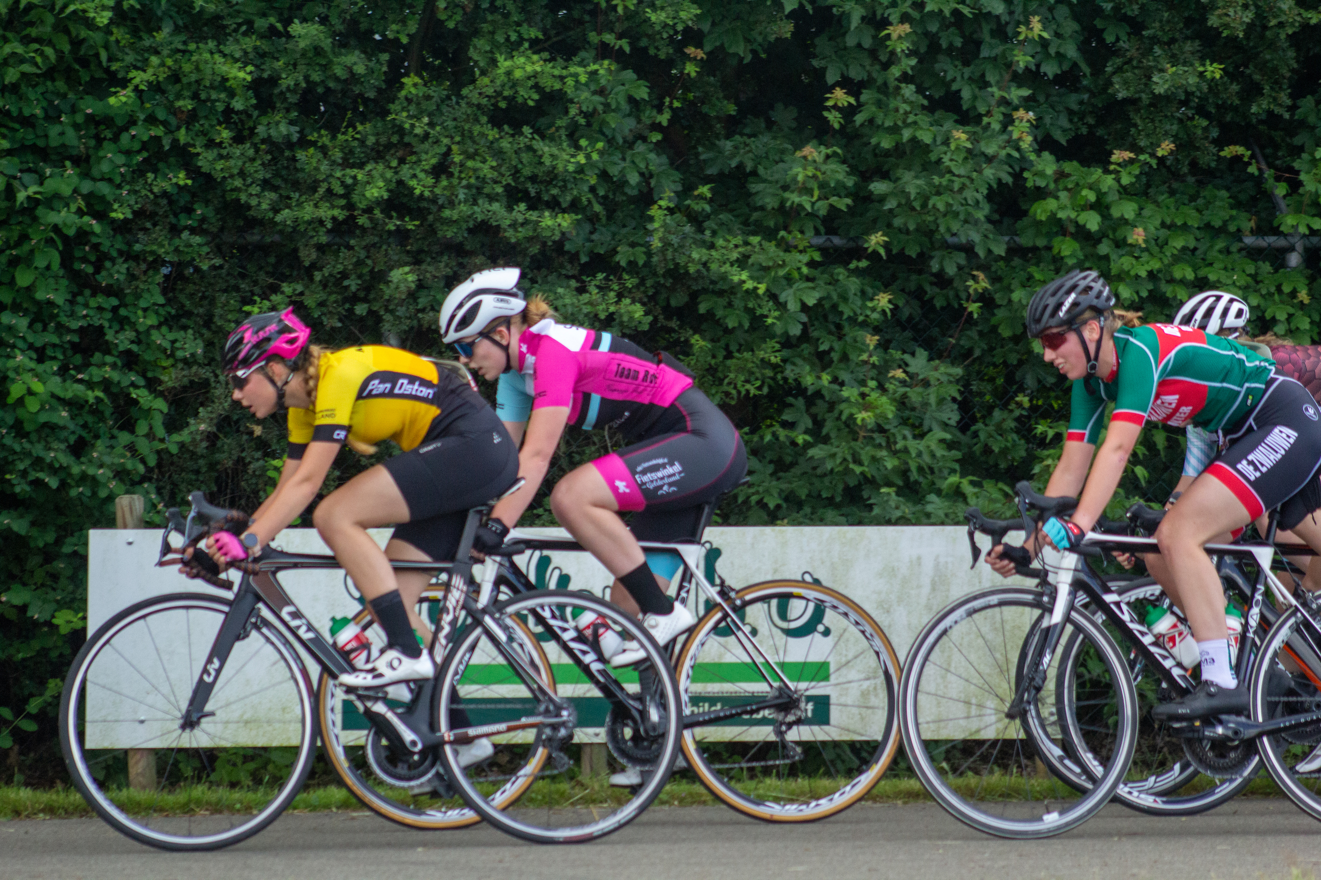 Three female cyclists racing on the road with one wearing a pink jersey and another wearing a yellow jersey.