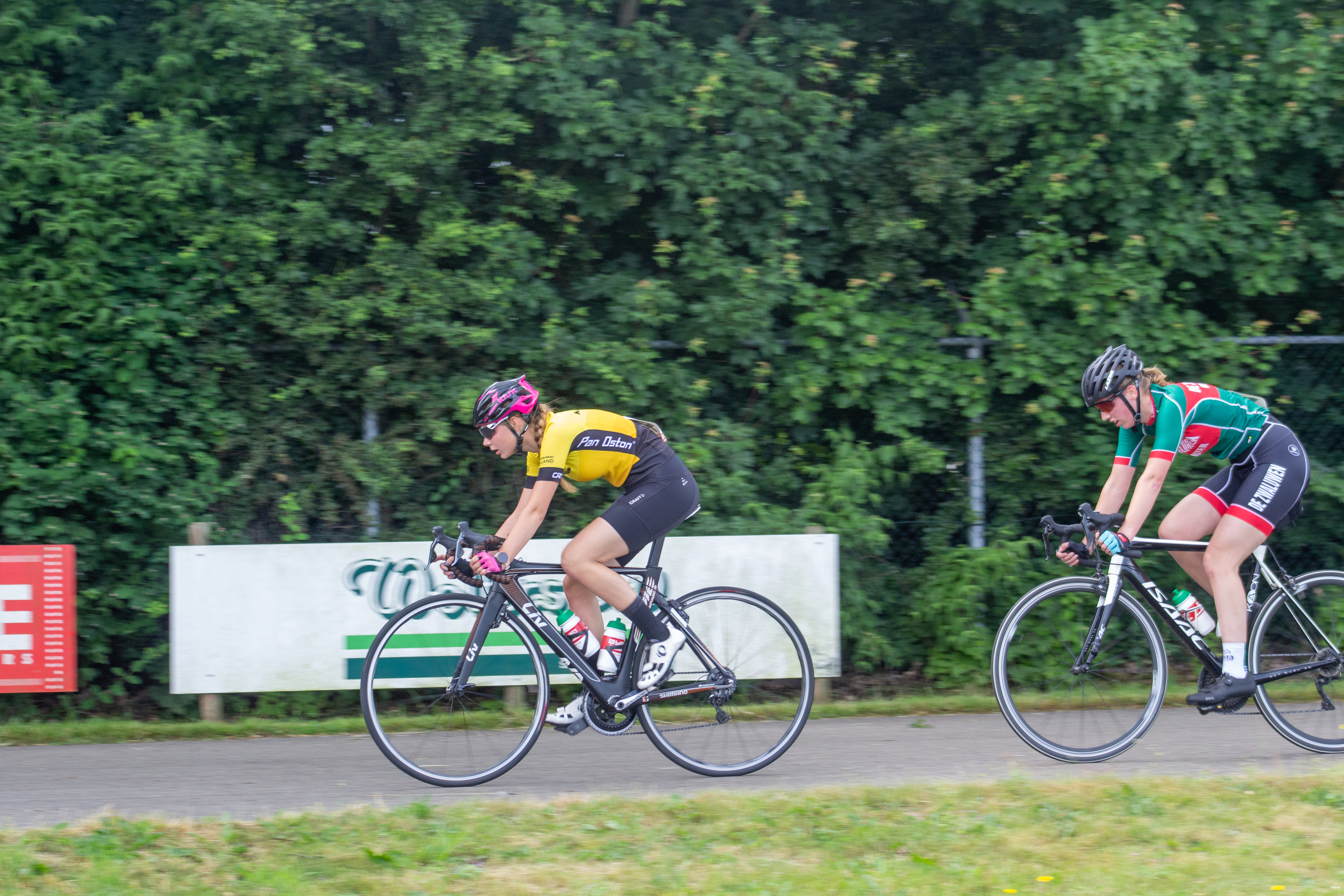 Two cyclists riding on a street with the words "Dames ronde van Deventer" in the background.