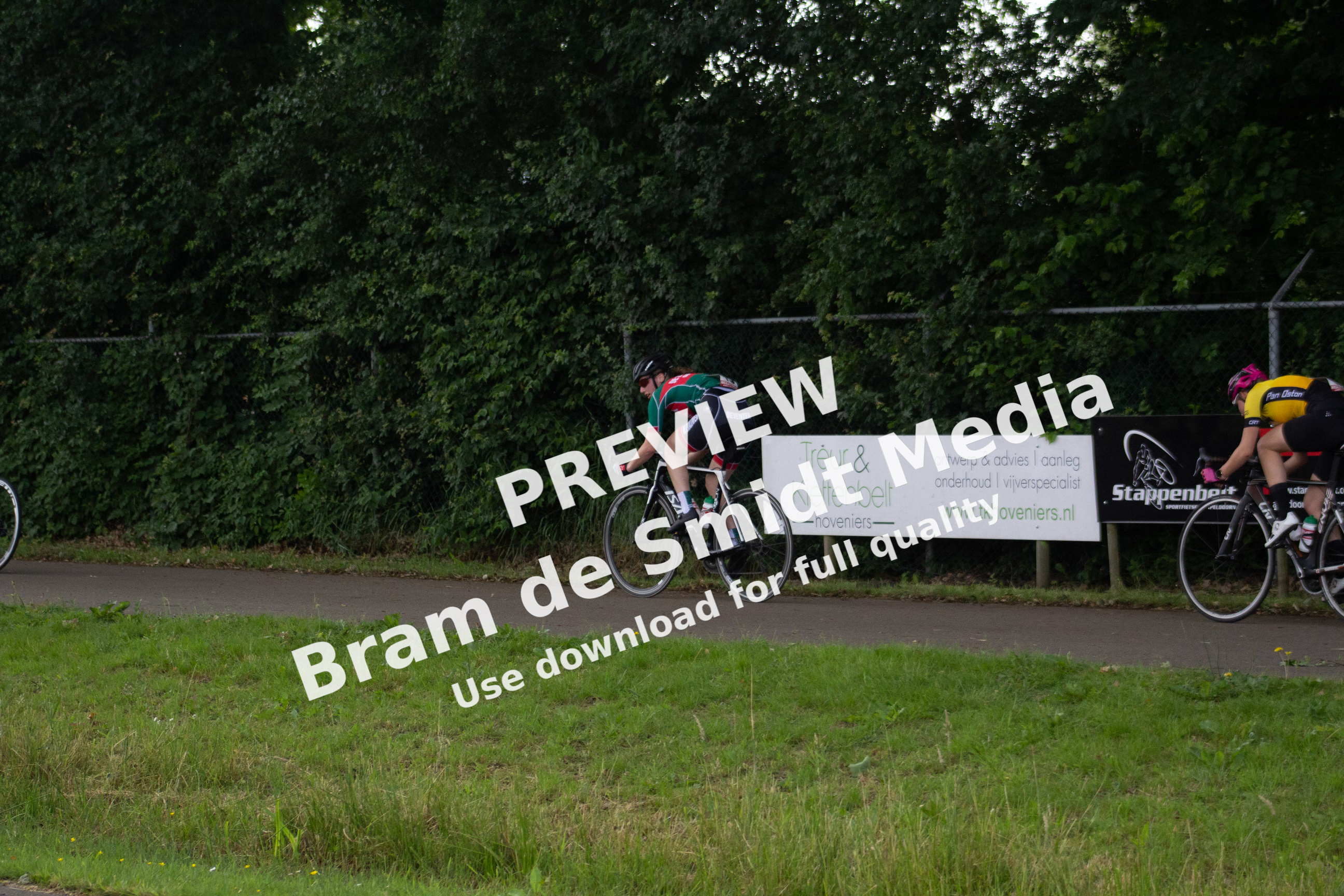 Two cyclists riding their bikes on a road with trees in the background and a banner that says Dames ronde van Deventer.