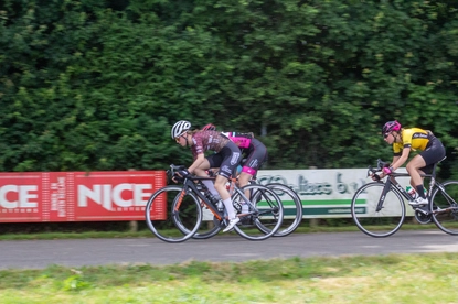 Three cyclists racing down a road near hedges and a red banner with the word "NICE".