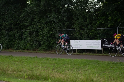 Two cyclists riding their bikes on a road with trees in the background and a banner that says Dames ronde van Deventer.