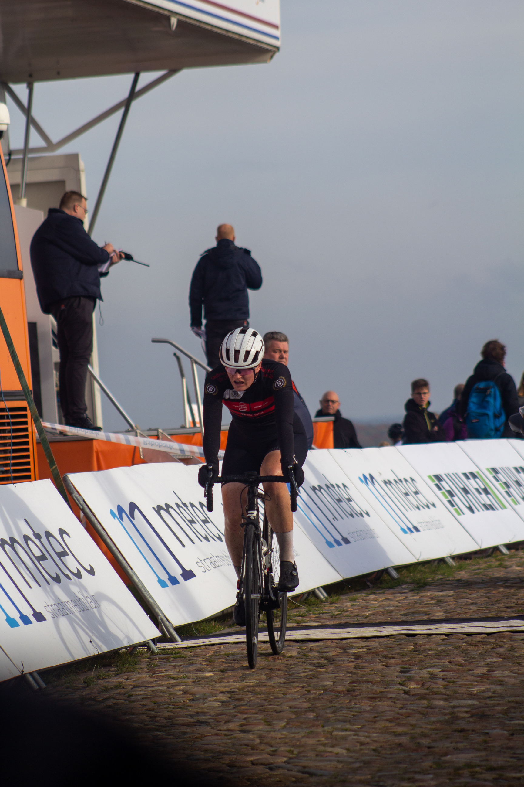 A person in a black and red cycling uniform on a cobblestone road near a barrier with the words NK Nieuweling Dames on it.