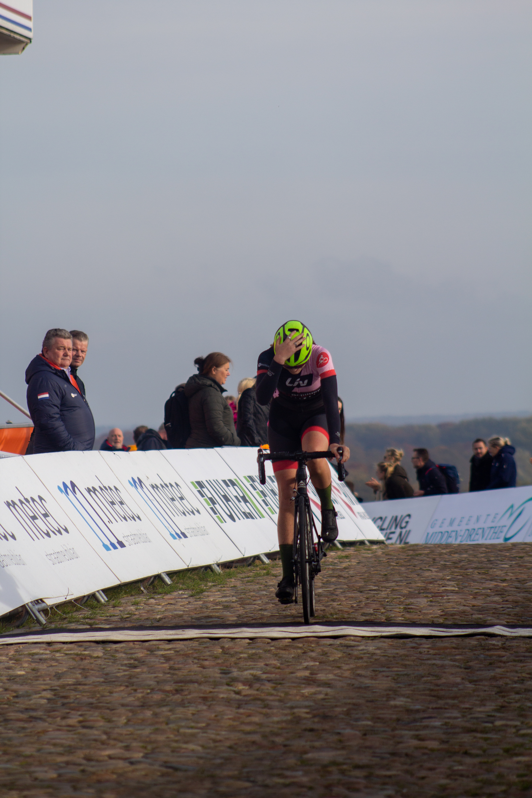 A cyclist wearing a yellow helmet and a pink and black jersey rides her bicycle down the road.