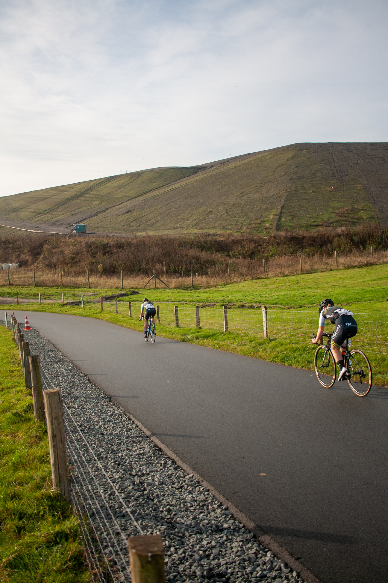 Two cyclists riding on a road near grass and a fence with one of them wearing a number 1 jersey.