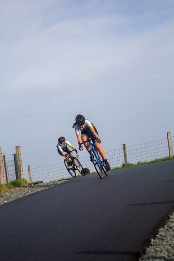 Two women are riding bikes in the same direction on a paved road.