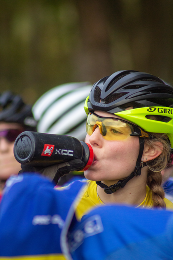 Two women wearing helmets are participating in a cycling race. One of them is drinking water from a KCC water bottle.