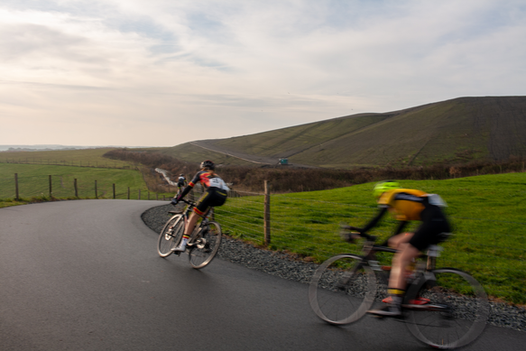 Two cyclists riding on a black paved road with one wearing the number 6.