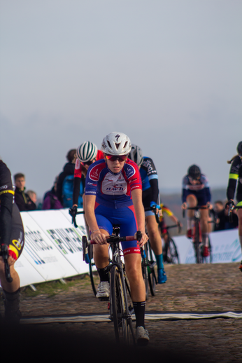 A cyclist wearing a blue Wielrennen shirt with the number 7 on it, jogs across a cobblestone road.