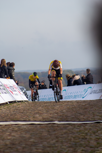 A group of cyclists ride along a cobblestone road near a banner for NK Nieuweling Dames.