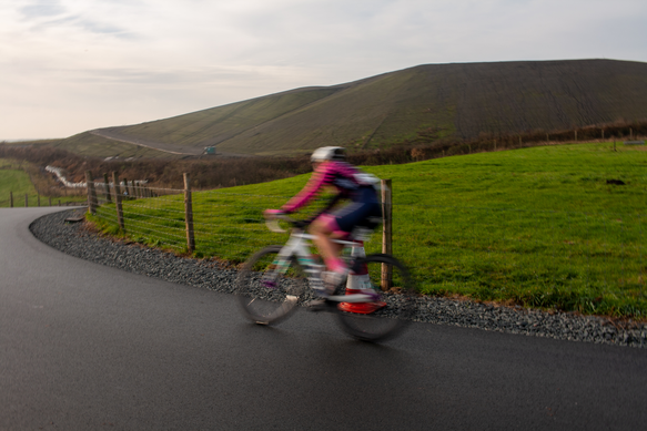 A woman in a pink jacket and helmet rides her bike on the side of a road.