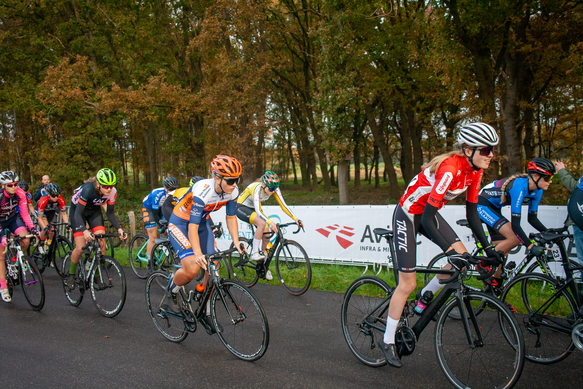 A group of cyclists racing down a street with signs behind them for NK Nieuweling Dames.