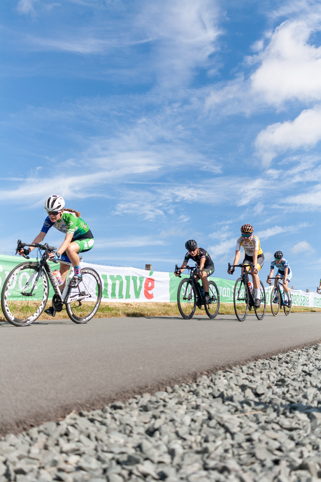 Four cyclists riding on a road, with the brand "Wielrennen" in their background.